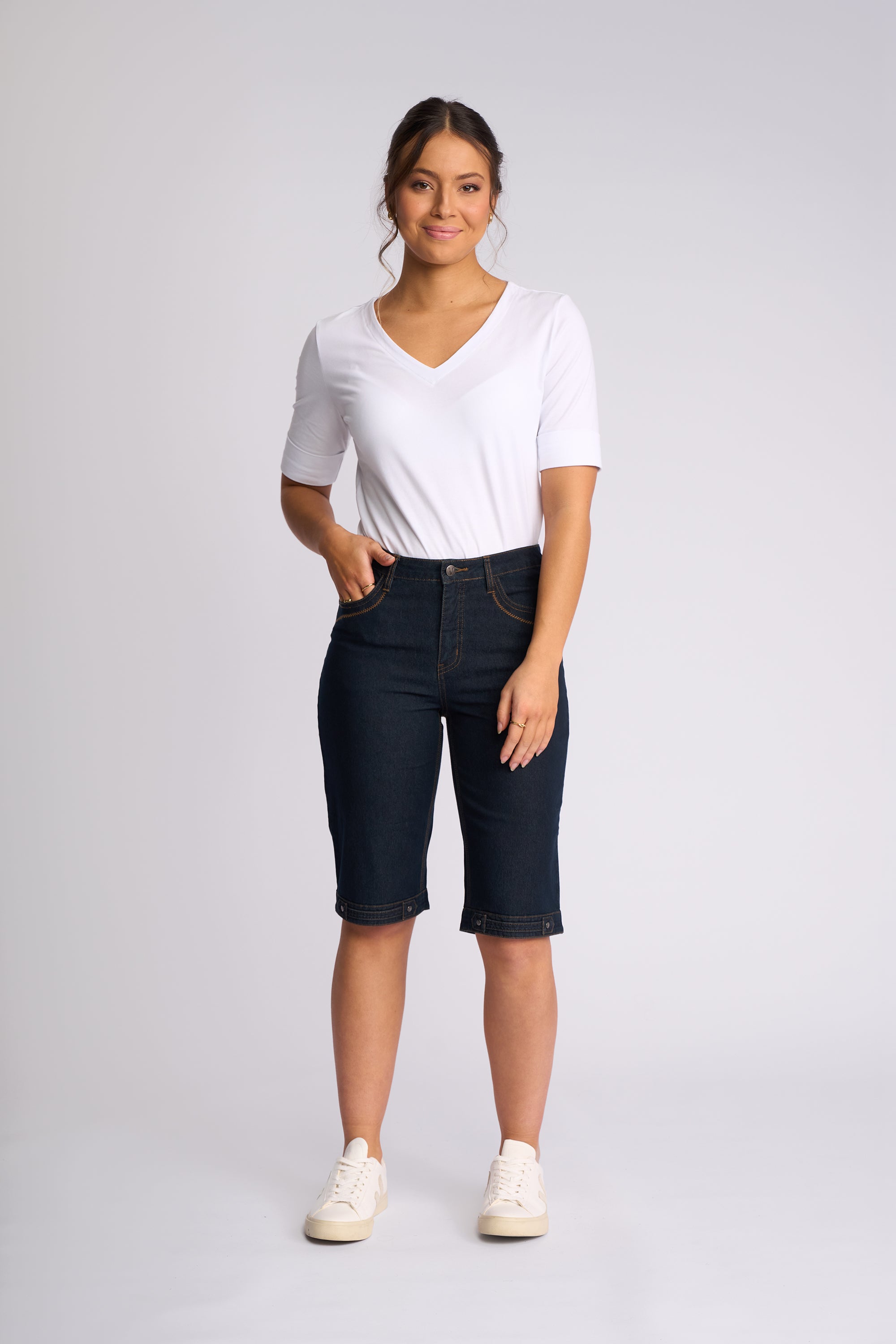 A woman modeling a pair of dark wash denim capri pants with a white V-neck t-shirt and white sneakers, standing against a light gray studio background.