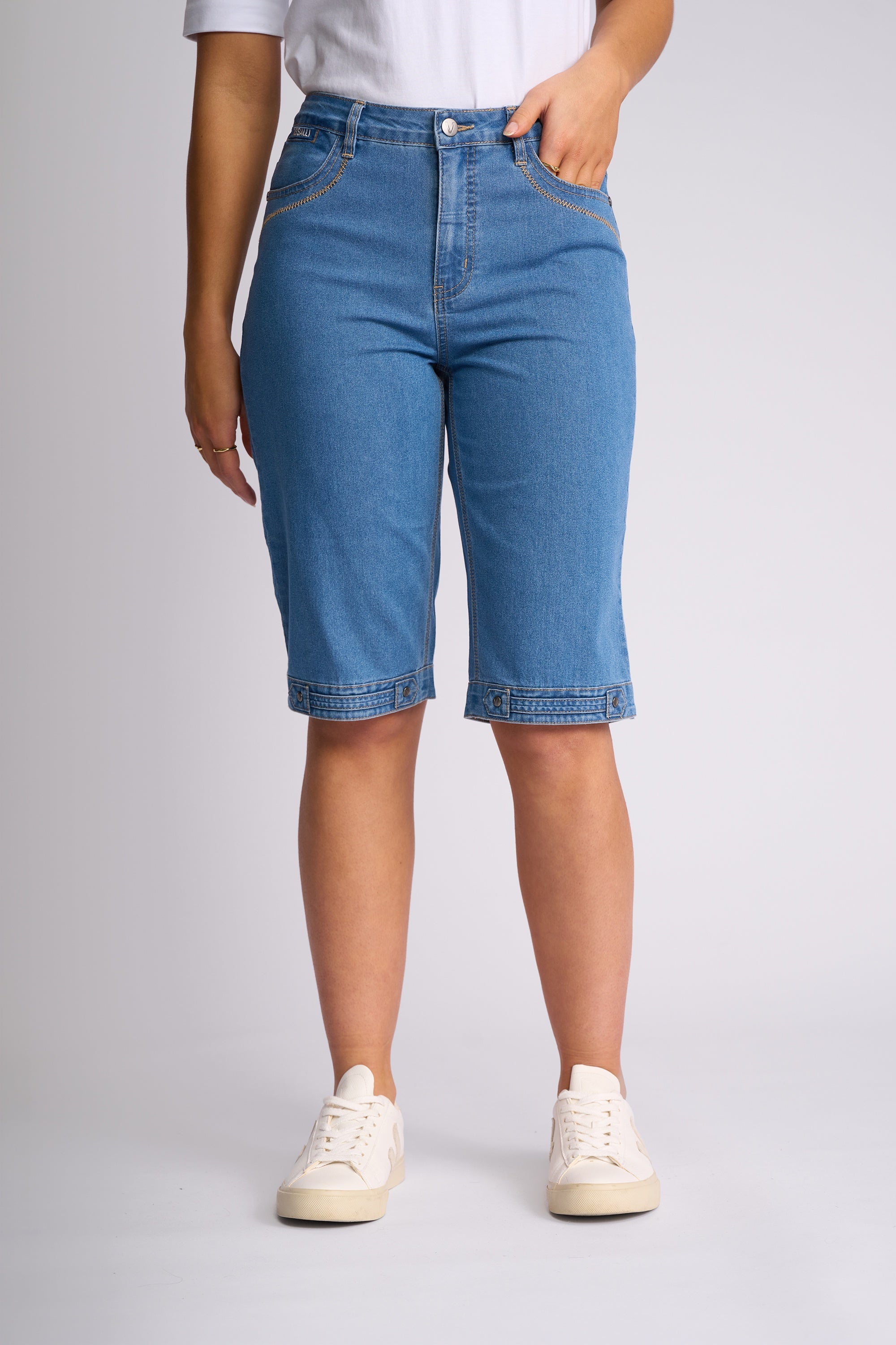 A model wearing a pair of high-waisted, light blue denim capri shorts with rolled hems, paired with a white t-shirt and white sneakers, shown from the waist down against a light gray studio background.