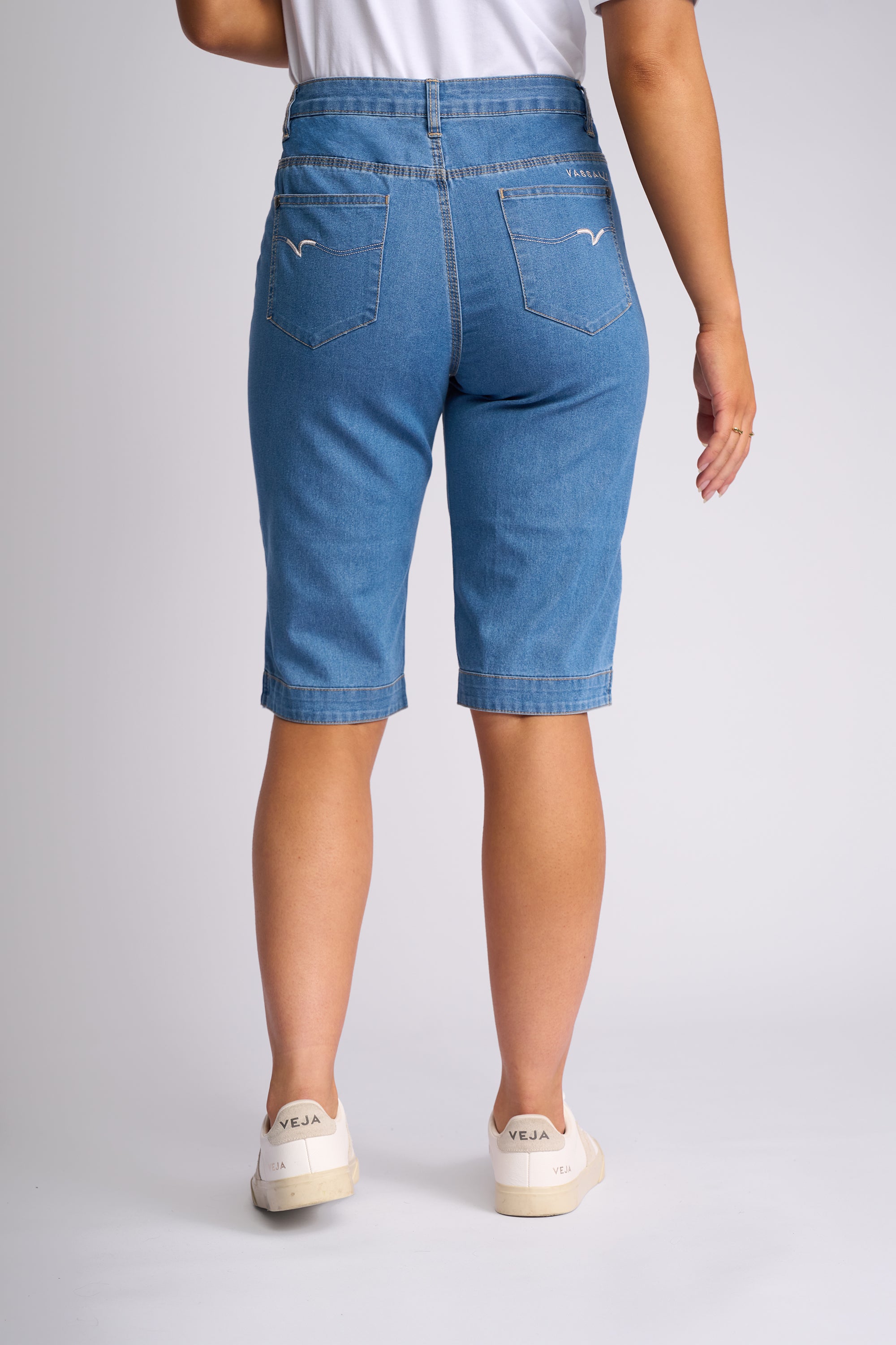 A person shown from the back wearing light blue denim capri shorts, a white top, and white slip-on shoes against a light gray studio background.