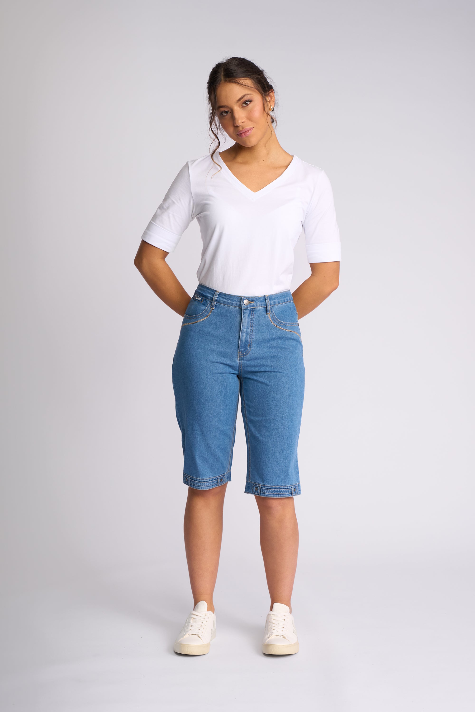 A woman modeling a white short-sleeve V-neck t-shirt and light blue denim capri jeans with white sneakers, standing against a light gray studio background.
