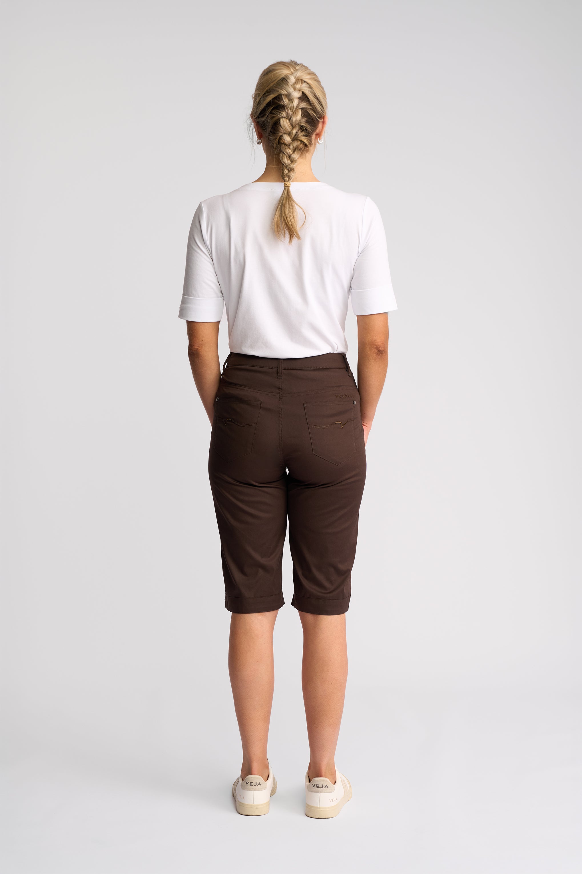 A woman viewed from behind wearing a white short-sleeve t-shirt, dark brown cropped shorts, and off-white sneakers, standing against a light gray studio background.