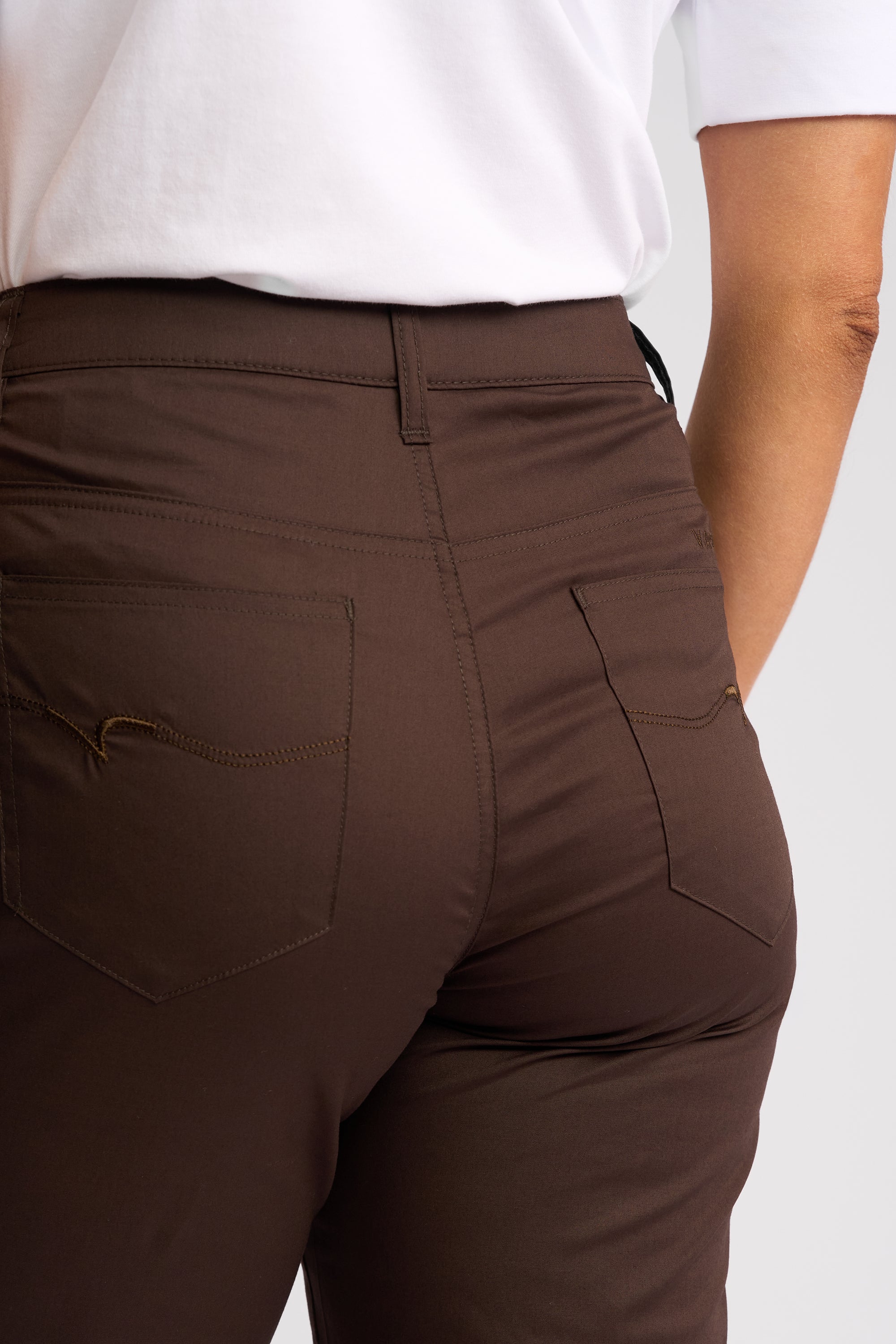 Close-up back view of a person wearing dark brown tailored pants with a white top, showing the back pockets and waistband.