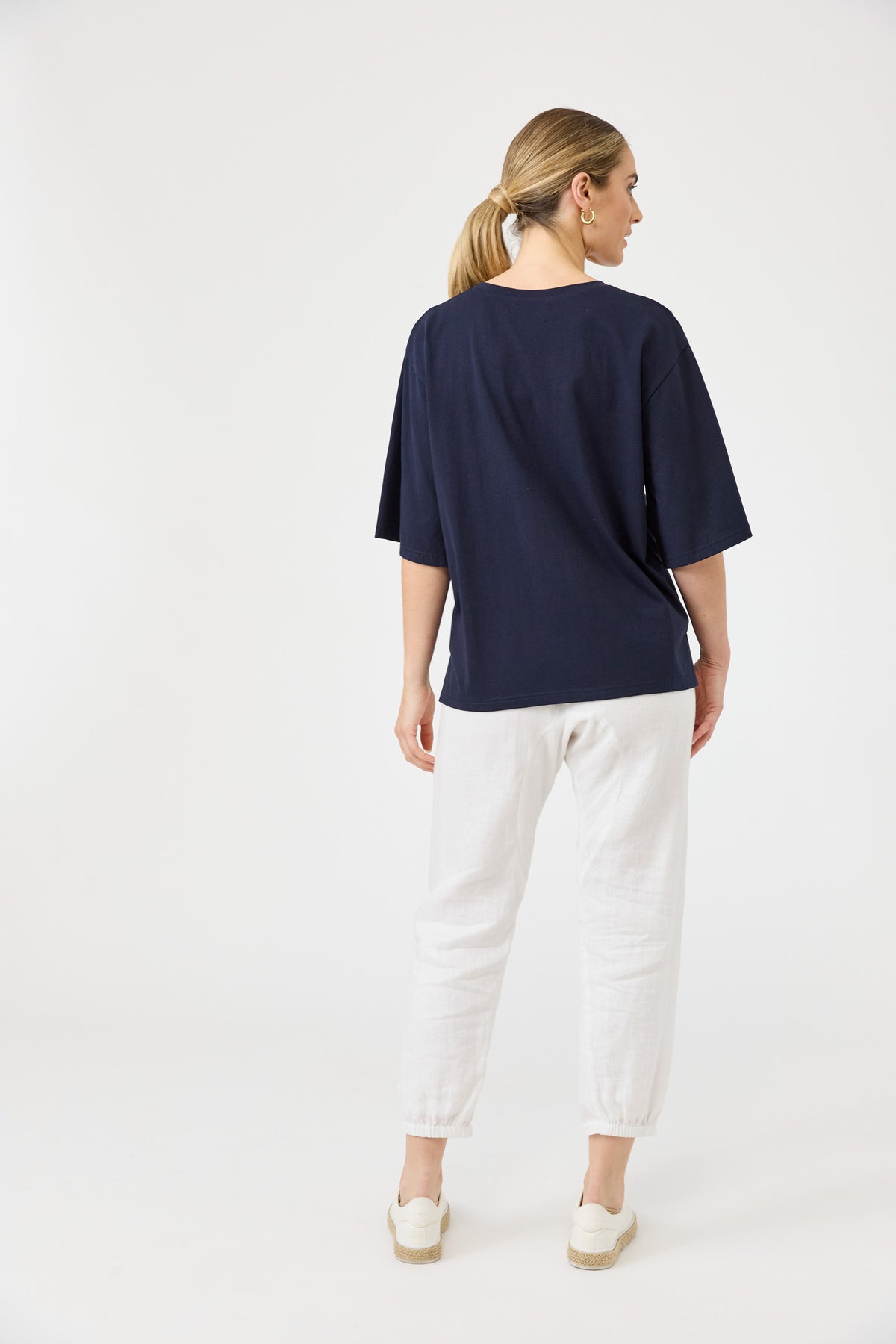 A woman viewed from the back wearing an oversized navy blue short-sleeve t-shirt, white cropped trousers, and white slip-on shoes, set against a plain white studio background.