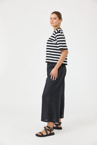 A woman modeling a black and white horizontally striped short-sleeve top paired with dark wide-leg pants and black sandals, standing against a plain white studio background.