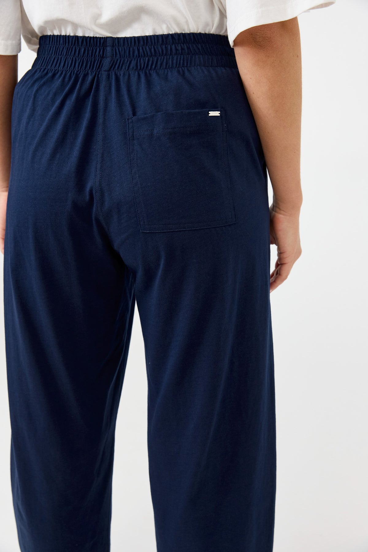Close-up back view of a person wearing dark navy drawstring pants with a rear patch pocket, paired with a white top against a plain light background.