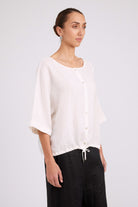 A woman wearing a loose white short-sleeve button-front top and black drawstring pants, photographed against a plain light gray studio background.