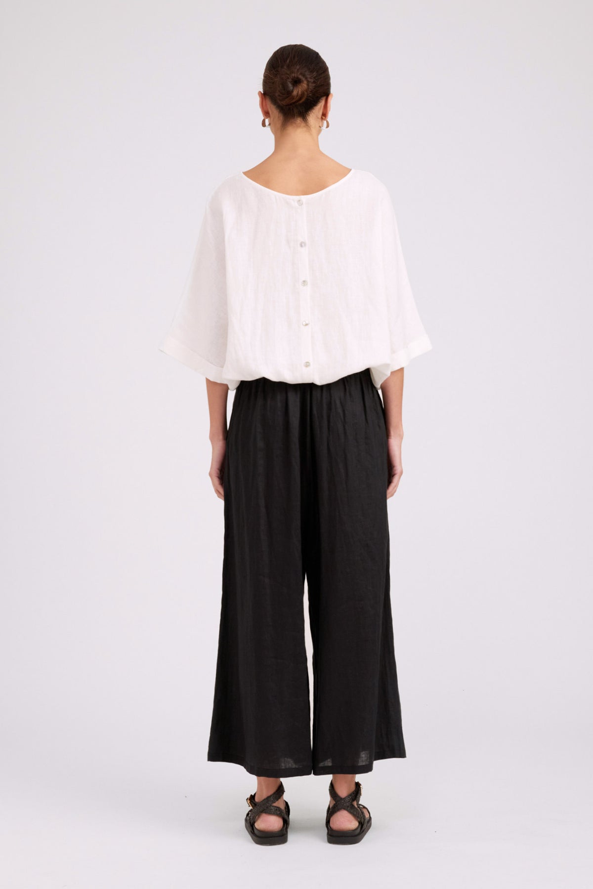 A woman shown from the back wearing a white short-sleeve button-front blouse and black wide-leg pants, standing against a light gray studio background.