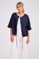 A woman modeling a navy blue short-sleeve button-front jacket over a white jumpsuit, photographed against a plain light gray studio background.