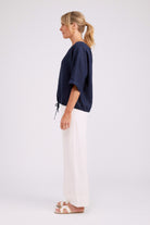 A woman shown in profile wearing a navy blue short-sleeve top with a drawstring and white wide-leg pants, standing against a plain light gray studio background.