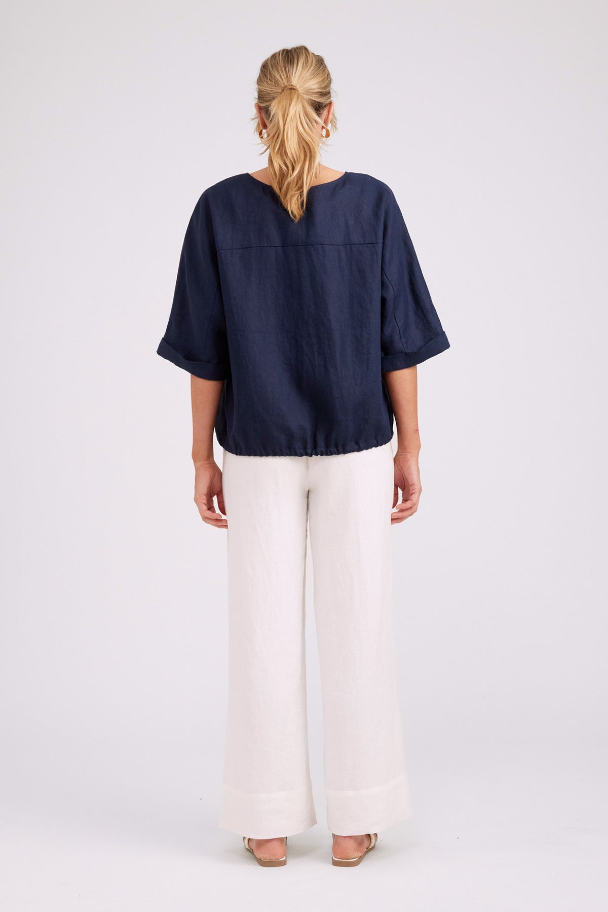 A woman viewed from the back wearing a navy blue short-sleeve top and white wide-leg pants against a light gray studio background.