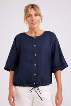 A woman wearing a navy blue short-sleeve linen top with a button-down front and drawstring hem, paired with white pants, against a light gray studio background.