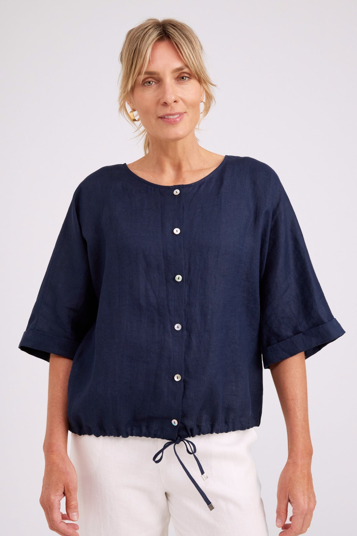 A woman wearing a navy blue short-sleeve linen top with a button-down front and drawstring hem, paired with white pants, against a light gray studio background.