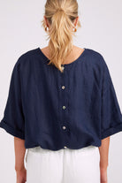 A woman shown from the back wearing a navy blue linen button-front top with rolled three-quarter sleeves and white pants, against a light gray studio background.