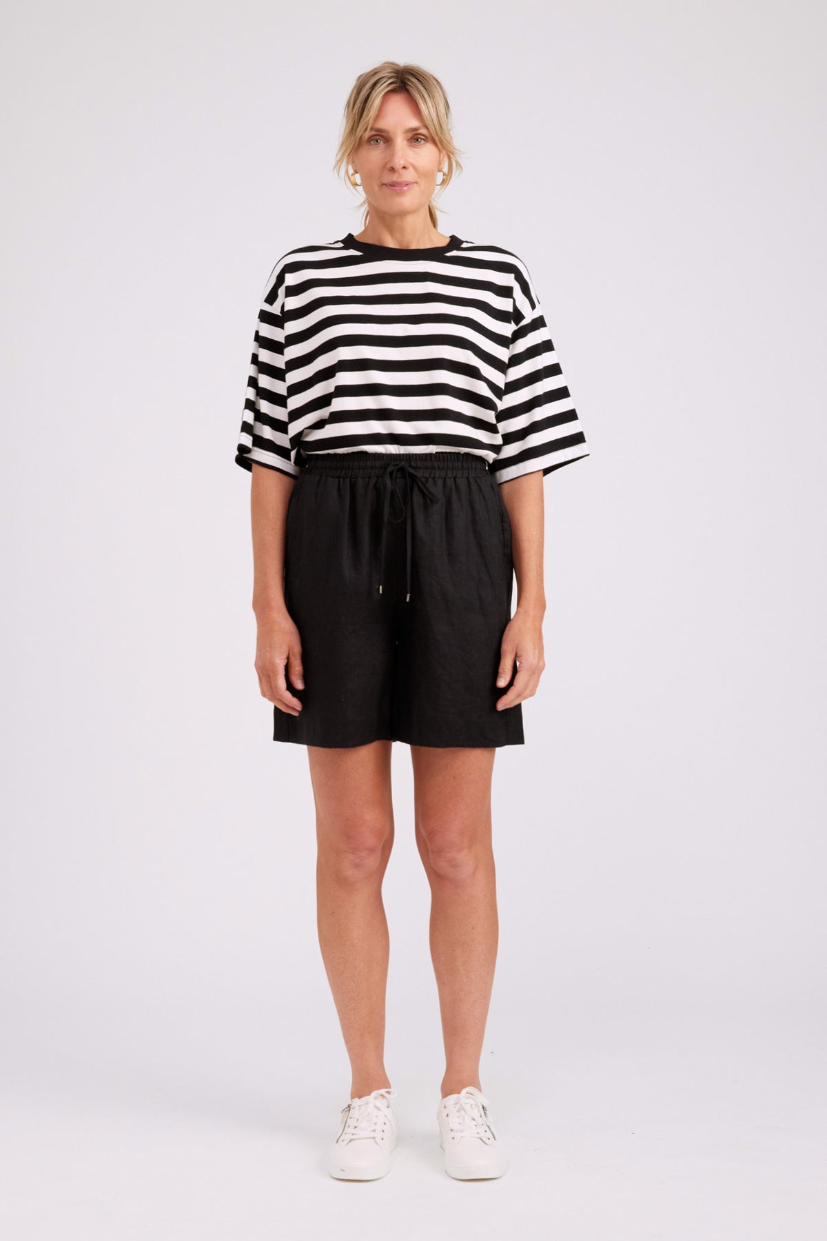 A woman modeling a black and white horizontally striped short-sleeve crop top with black drawstring shorts and white sneakers, standing against a light gray studio background.