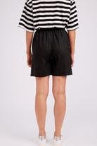 Rear view of a model wearing black high-waisted shorts, a black and white striped short-sleeve top, and white sneakers against a light gray studio background.