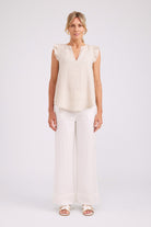 A woman modeling a light cream sleeveless top with ruffled shoulders and a split V-neck, paired with matching wide-leg trousers and white sandals, against a plain light gray studio background.