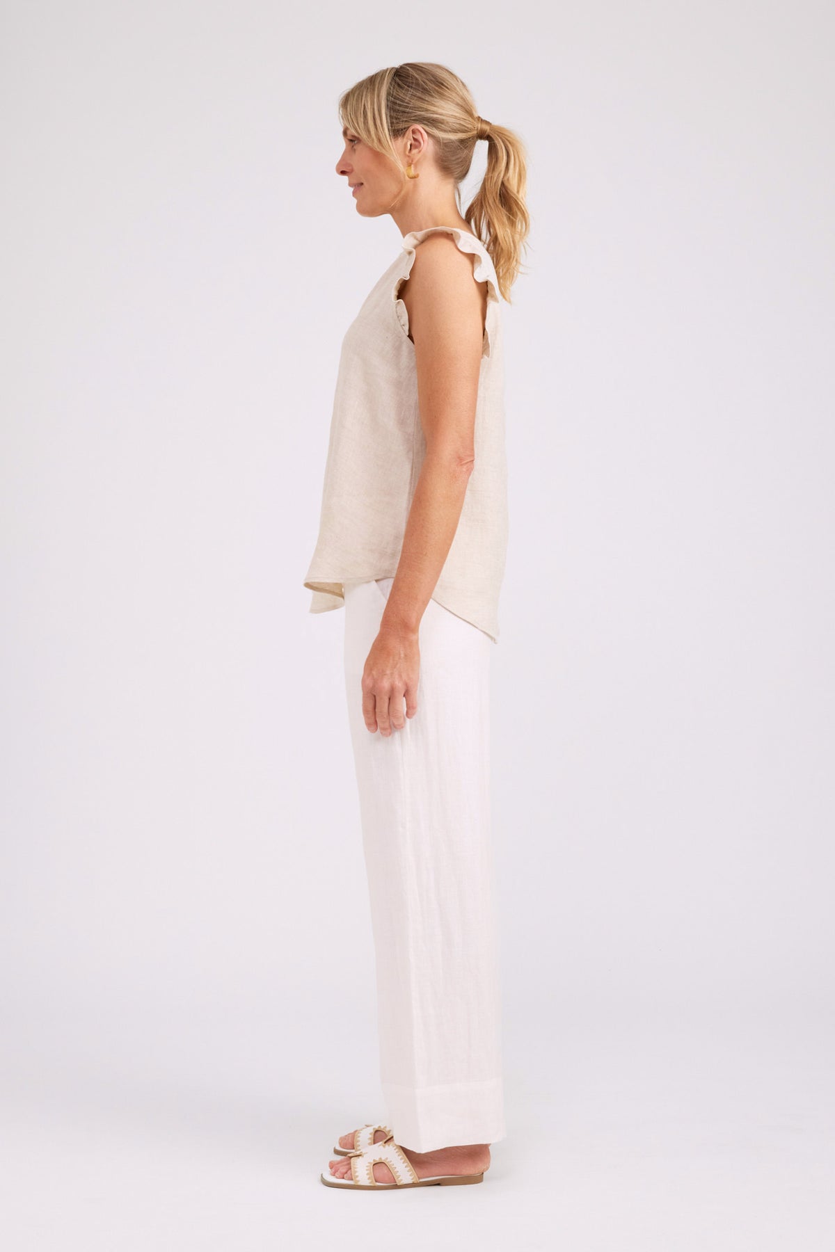 A woman shown in side profile wearing a sleeveless cream top and white wide-leg pants against a plain light gray studio background.