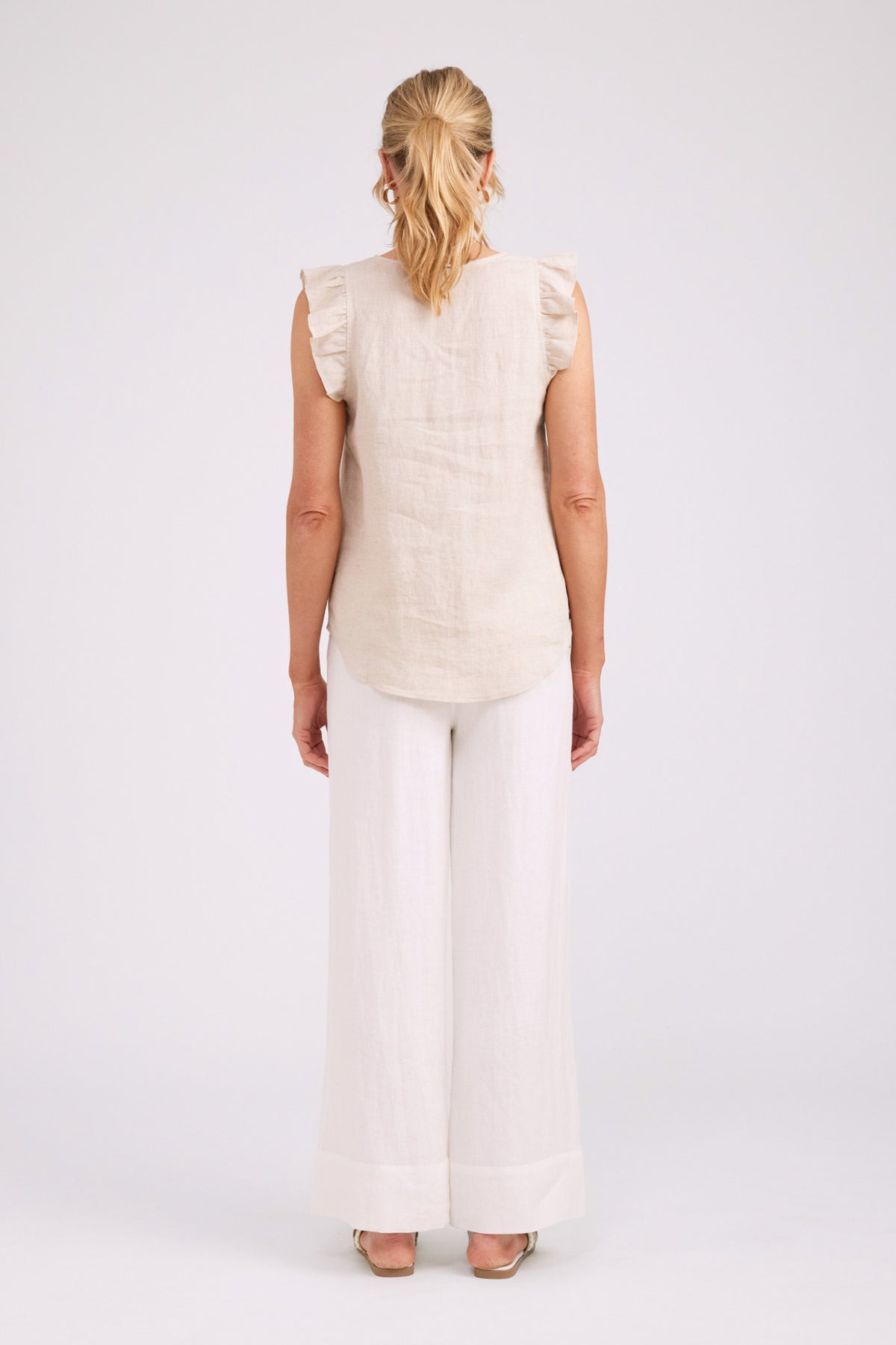 A woman viewed from the back wearing a light beige sleeveless top with ruffled shoulders and white wide-leg pants, standing against a plain light gray studio background.