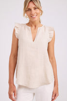 A woman wearing a sleeveless cream linen top with ruffled cap sleeves and a split neckline, paired with white pants, against a light gray studio background.