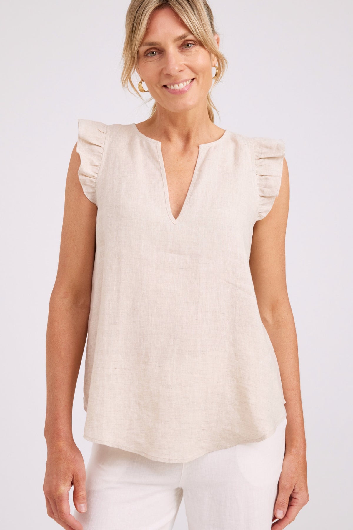 A woman wearing a sleeveless cream linen top with ruffled cap sleeves and a split neckline, paired with white pants, against a light gray studio background.