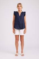 A woman modeling a sleeveless navy blue top with ruffled shoulders and a split V-neckline, paired with white shorts and white sandals, against a light gray studio background.