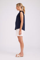 A woman shown in profile wearing a sleeveless navy top with ruffled shoulder details, white shorts, and white slide sandals, photographed against a light gray studio background.