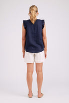A woman viewed from the back wearing a sleeveless navy top with ruffled shoulders and white shorts, standing against a light gray studio background.