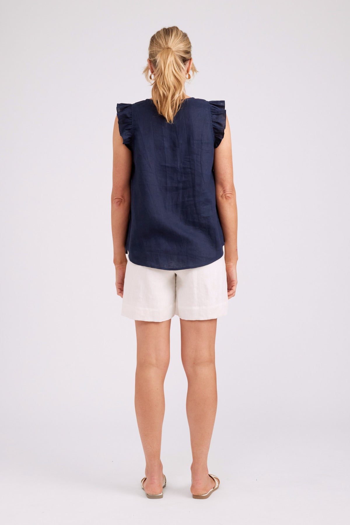 A woman viewed from the back wearing a sleeveless navy top with ruffled shoulders and white shorts, standing against a light gray studio background.