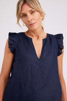 A woman wearing a navy blue sleeveless top with ruffled cap sleeves and a split V-neckline, photographed against a plain light gray studio background.