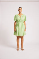 A woman modeling a light green textured mini dress with short flutter sleeves, a gathered waist, and a V-neckline, standing against a plain light gray studio background.