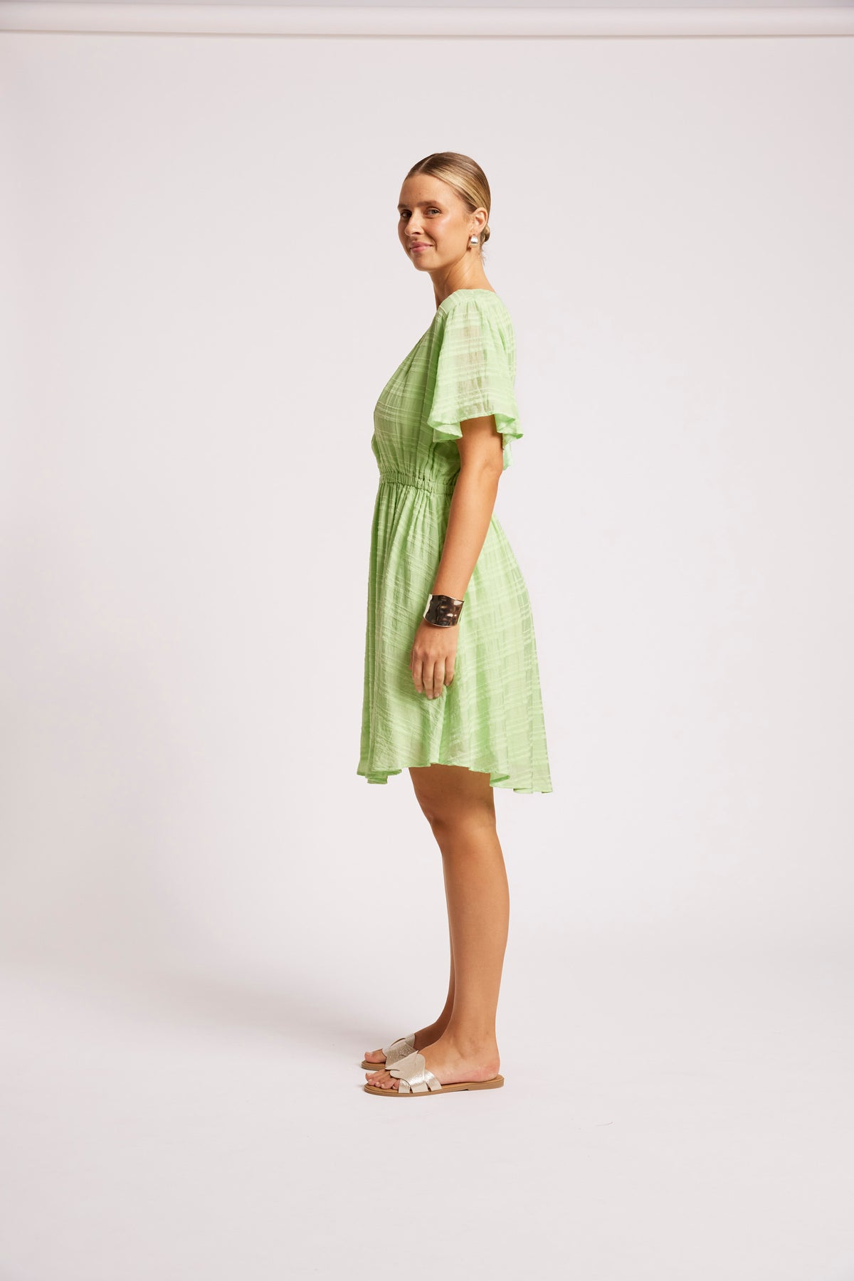 A woman modeling a light green textured mini dress with short flutter sleeves, standing in profile against a plain off-white studio background.