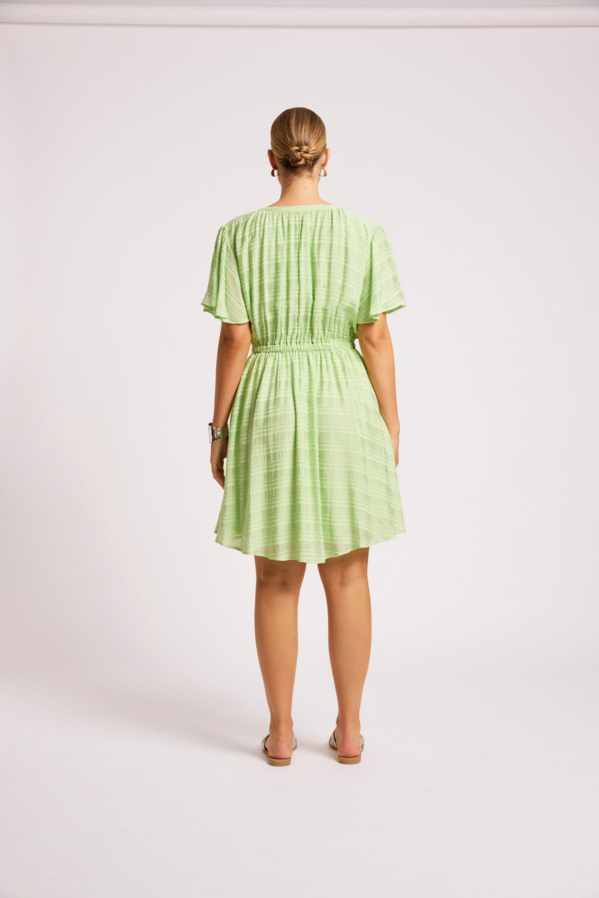 A woman viewed from the back wearing a light green pleated short-sleeve dress with a gathered waist, standing against a plain light gray studio background.