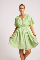 A woman modeling a light green textured mini dress with short flutter sleeves, a gathered waist, and a V-neckline, photographed against a plain light gray studio background.