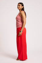 A woman stands in profile wearing a sleeveless red and white striped top and bright red wide-leg pants against a plain light gray studio background.