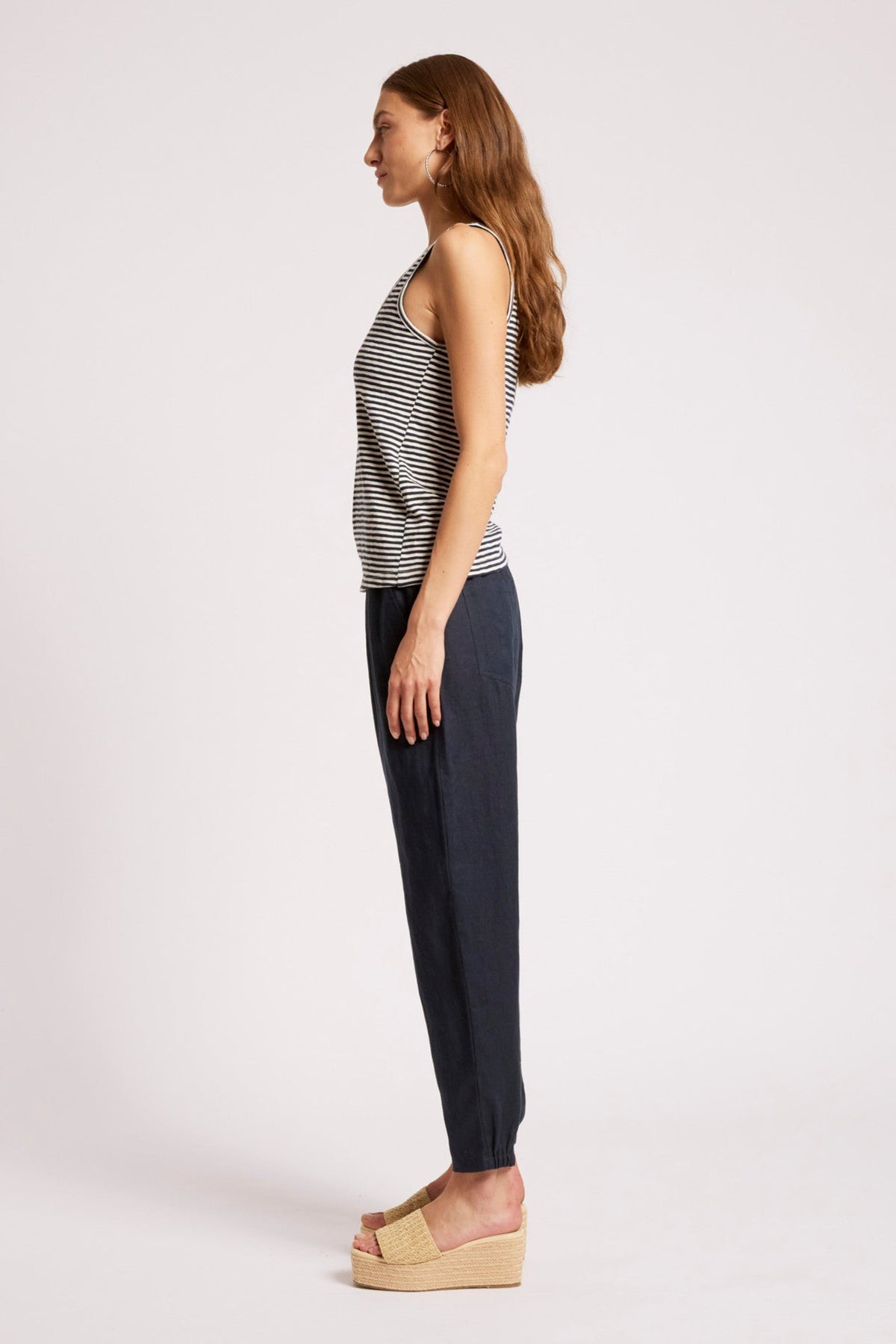 A woman shown in profile wearing a sleeveless black and white striped top, dark navy pants, and beige platform sandals against a plain light gray studio background.