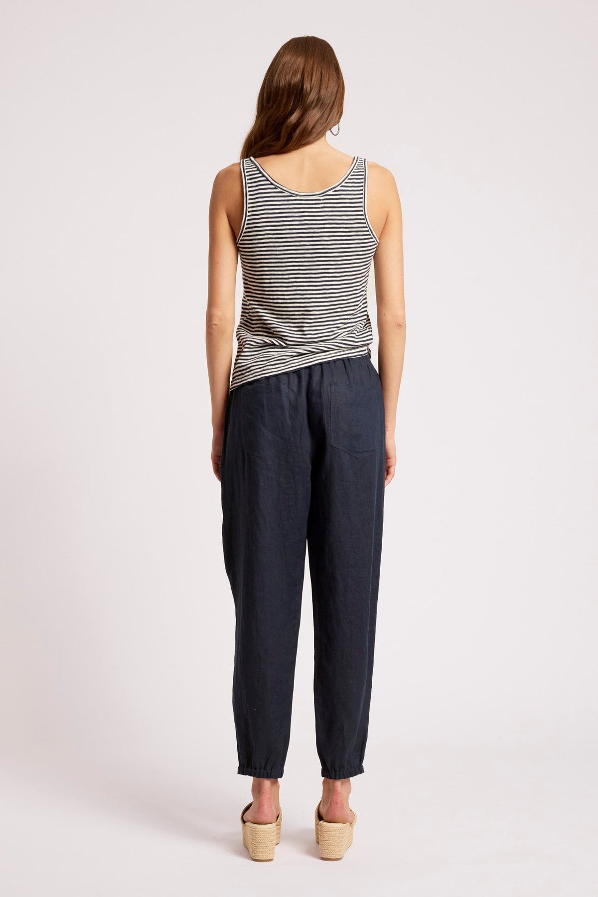 A woman shown from the back wearing a sleeveless black and white striped top and dark navy pants, standing against a plain light gray studio background.