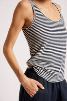 A close-up of a model wearing a navy and white horizontally striped tank top, paired with dark navy pants, against a plain light gray background.