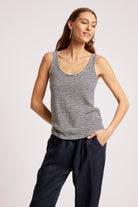 A woman modeling a sleeveless tank top with thin black and white horizontal stripes, paired with dark navy pants, against a plain light gray studio background.