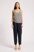 A woman modeling a sleeveless top with thin black and white horizontal stripes, paired with dark navy blue pants and beige platform sandals, against a light gray studio background.