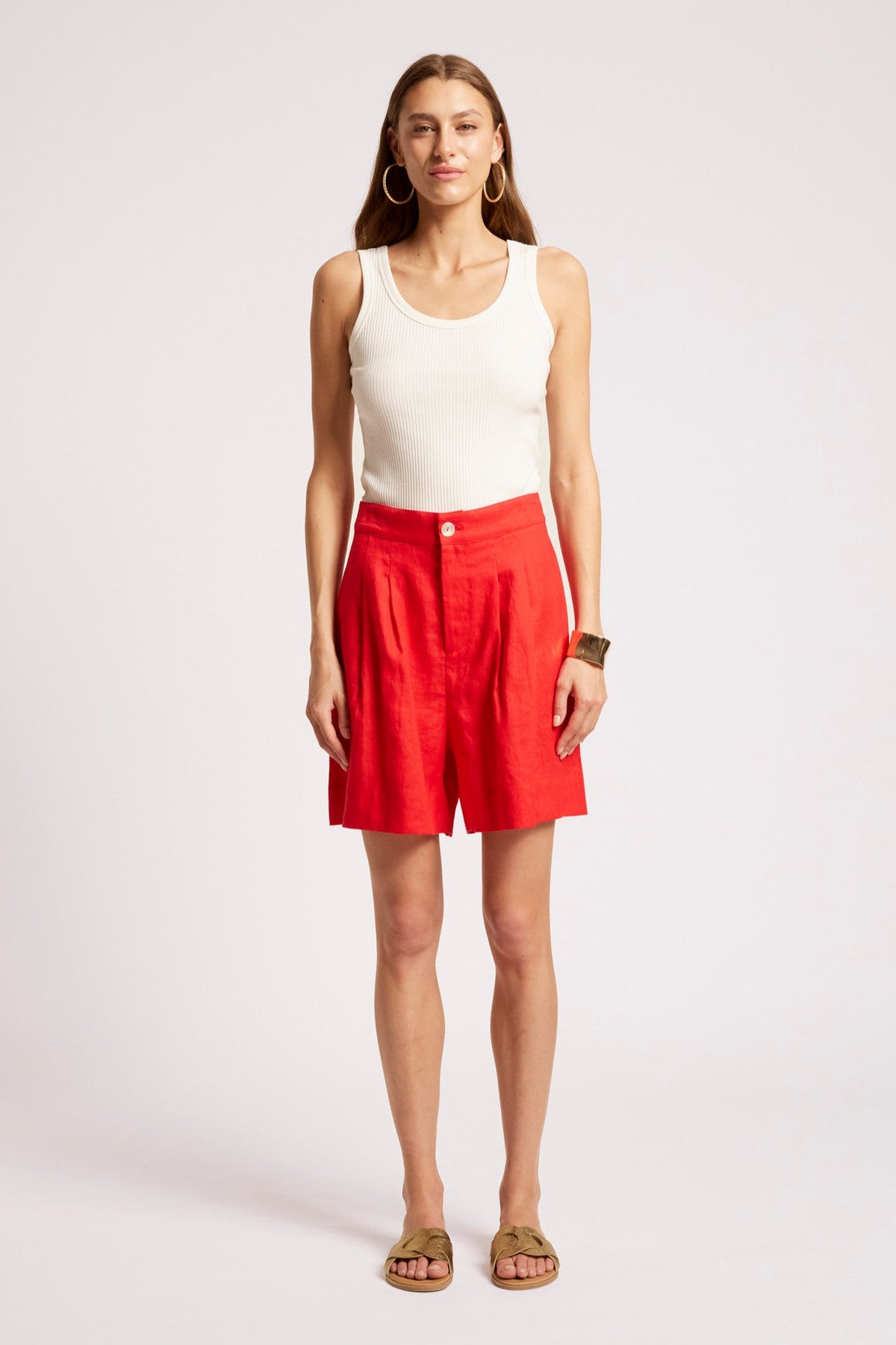 A woman modeling bright red high-waisted shorts with a white ribbed tank top and tan slide sandals, standing against a light gray studio background.