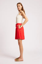 A woman modeling a bright red wrap-style mini skirt with a matching red belt, paired with a white halter top and flat sandals, against a plain light gray studio background.