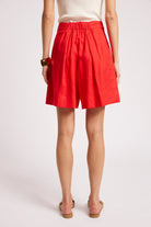 A model from the back wearing bright red pleated shorts, a cream ribbed top, and tan flat sandals against a light gray studio background.
