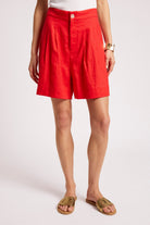A model wearing bright red linen shorts with pleats, styled with a white top and brown slide sandals against a light gray studio background.
