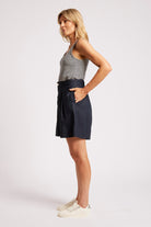 A woman modeling a pair of dark navy blue high-waisted shorts with a striped tank top and white sneakers, standing against a light gray studio background.