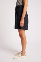 A side-profile view of a model wearing a dark navy blue pleated linen-blend mini skirt, paired with a black and white striped top and white sneakers, against a plain light gray studio background.