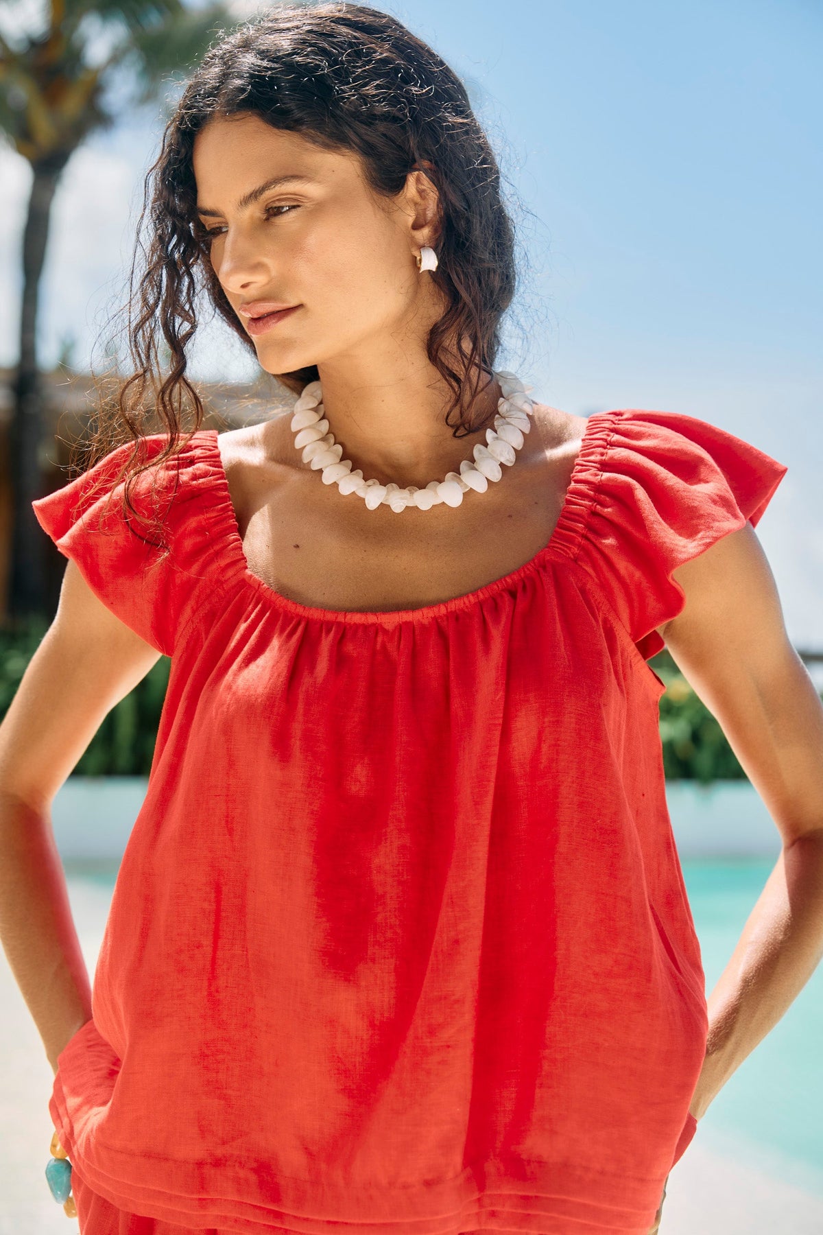 A woman wearing a bright red off-the-shoulder top with ruffled sleeves, styled with a chunky white shell necklace, photographed outdoors near a pool with palm trees in the background.