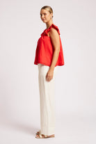 A woman modeling a bright red sleeveless top with a draped collar and white wide-leg pants against a plain light gray studio background.