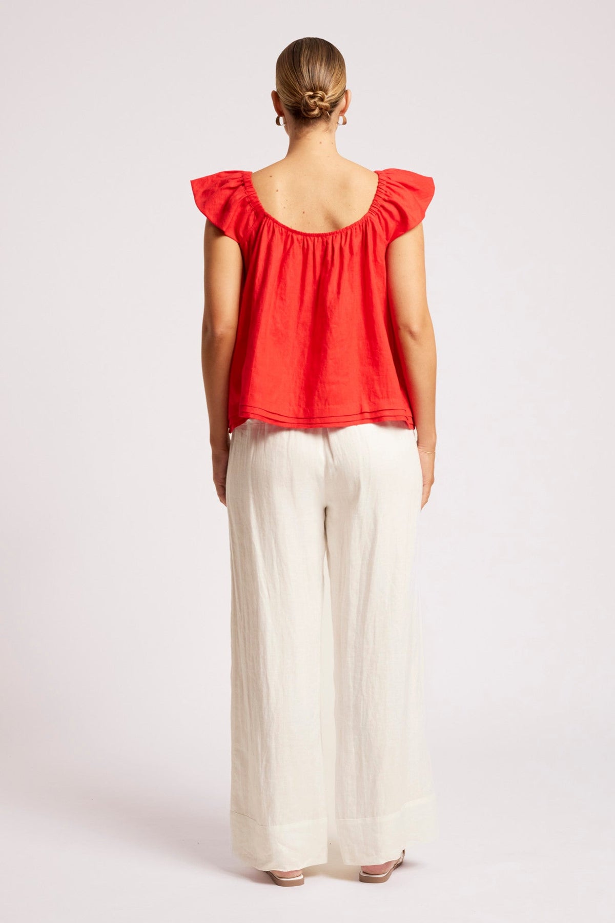 A woman viewed from the back wearing a bright red ruffled-sleeve top and off-white wide-leg pants, standing against a plain light gray studio background.