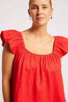 A woman wearing a bright red linen top with ruffled cap sleeves and a square neckline, photographed against a light gray studio background.