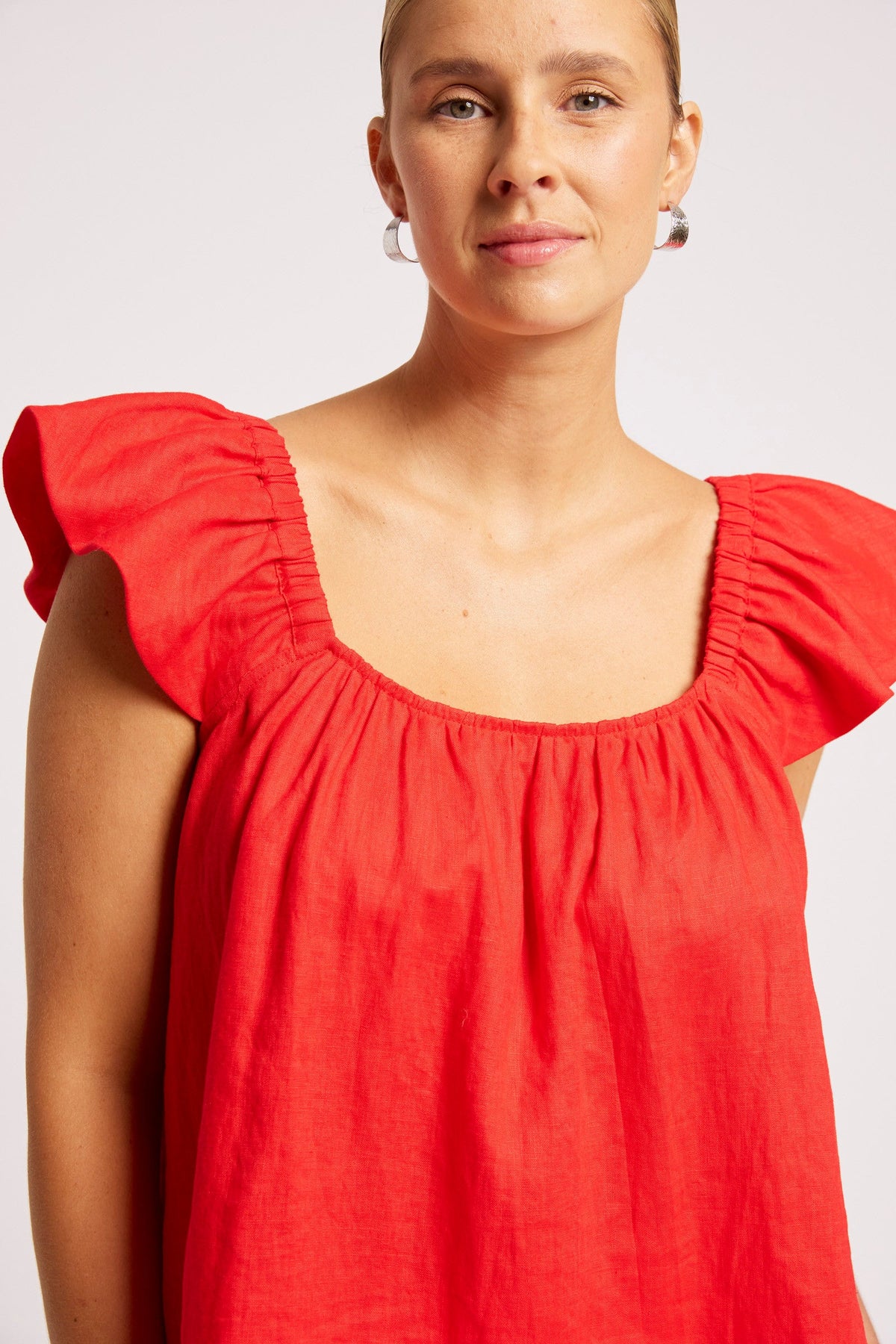 A woman wearing a bright red linen top with ruffled cap sleeves and a square neckline, photographed against a light gray studio background.